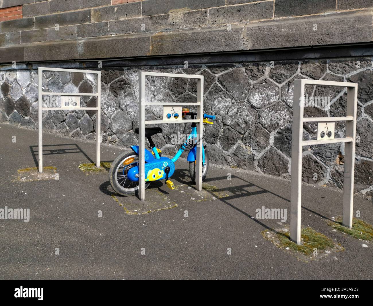 A vibrant blue child's bicycle is parked securely between two metal bike racks on a stone wall-lined street. - Smartphone Captured Stock Image