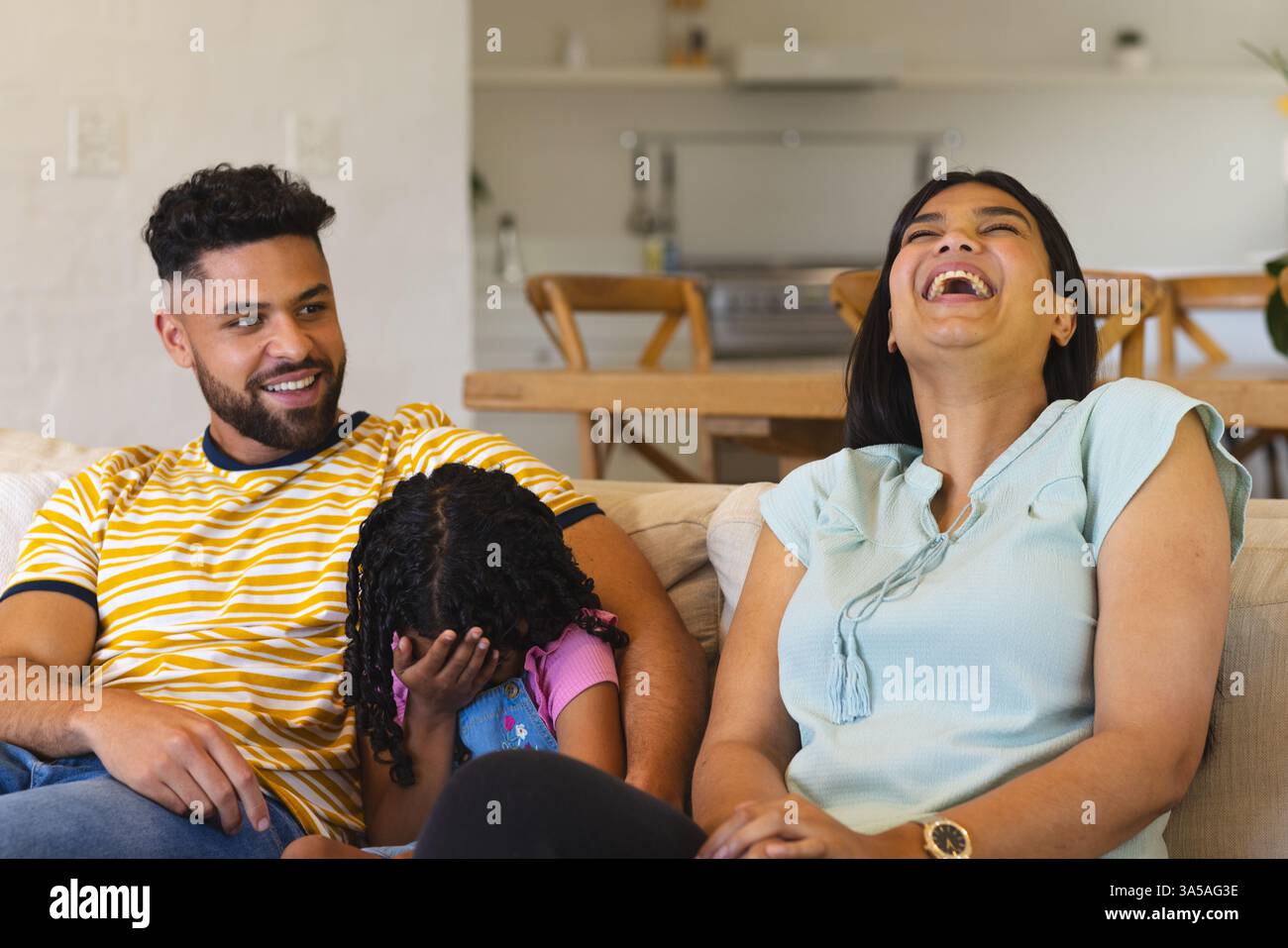 Happy family relaxing on couch, mother laughing while daughter playfully covers face Stock Photo