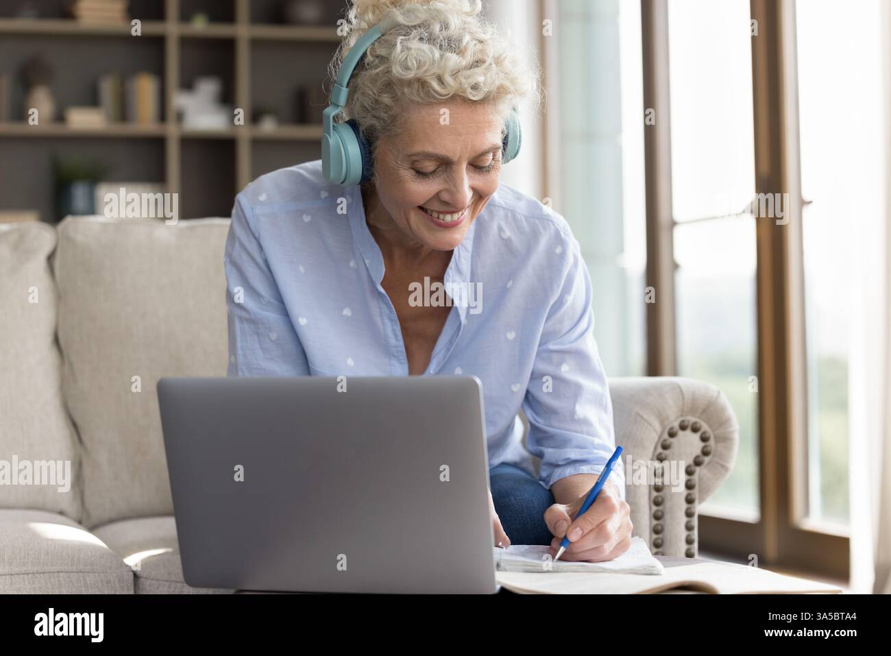 Elderly lady in headset watching webinar on notebook taking notes Stock Photo