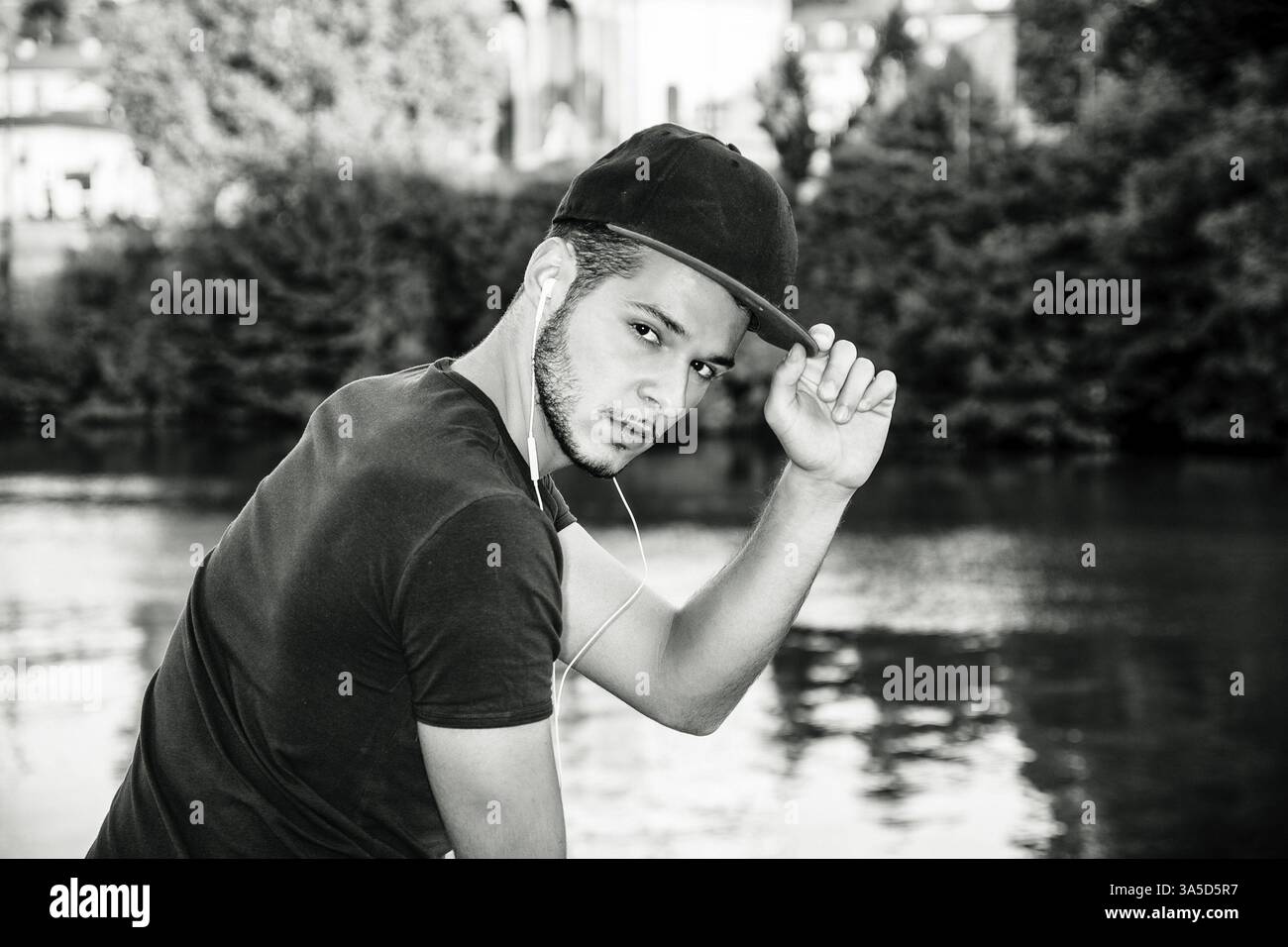 Portrait of contemplative light brown haired young man wearing baeball cap, beside picturesque river or lake, looking at camera Stock Photo