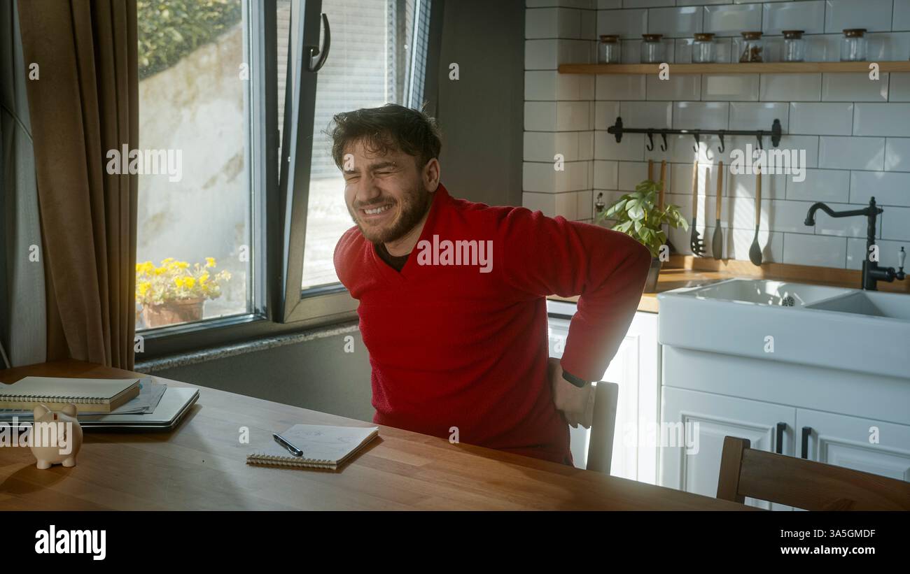 Man Experiencing Back Pain While Sitting in a Cozy Kitchen Stock Photo