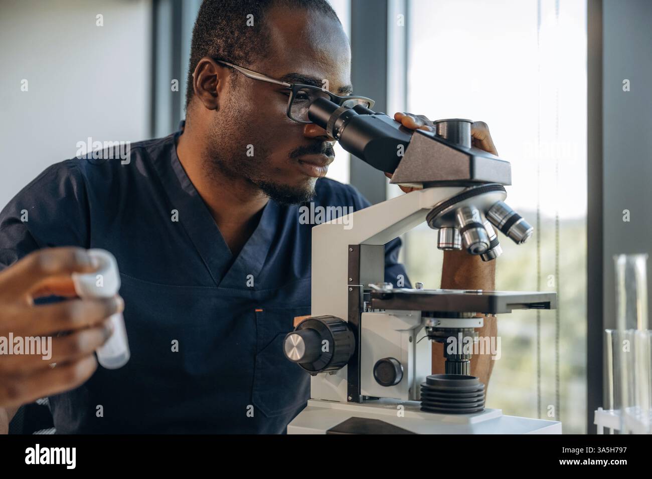 Test tube in hand, using the microscope. Scientist is working indoors. Stock Photo