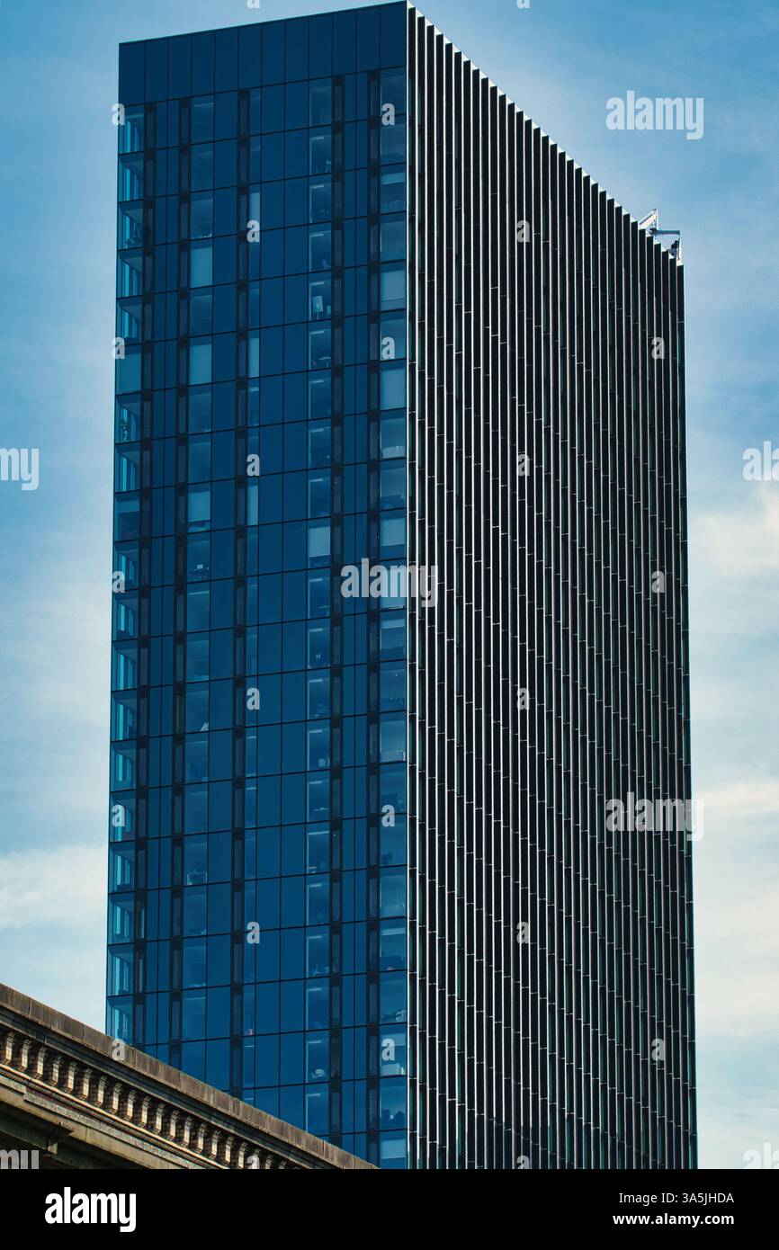 A tall building with blue glass and vertical metal slats under a cloudy sky. The bottom of another building is visible in the lower corner in Manchest Stock Photo