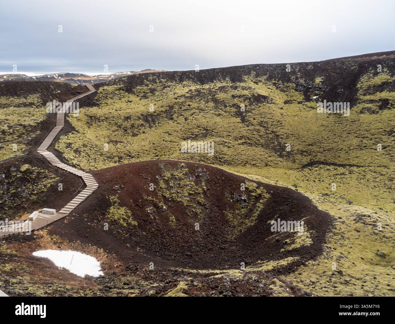 Stóra Grábrók, a volcanic crater in Iceland, features a wooden path winding up its moss-covered slopes. A surreal blend of red volcanic rock and green Stock Photo
