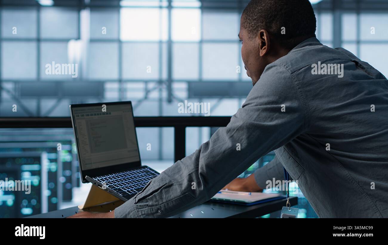 Technician coding on laptop in data center, managing server virtualization, finetuning network protocols. Server farm employee programming on notebook, enforcing advanced security measures, camera A Stock Photo