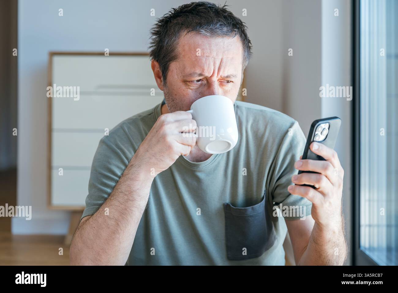 Drinking coffee and surfing the net on smartphone at home, portrait of mature adult male during his morning routine, selective focus Stock Photo