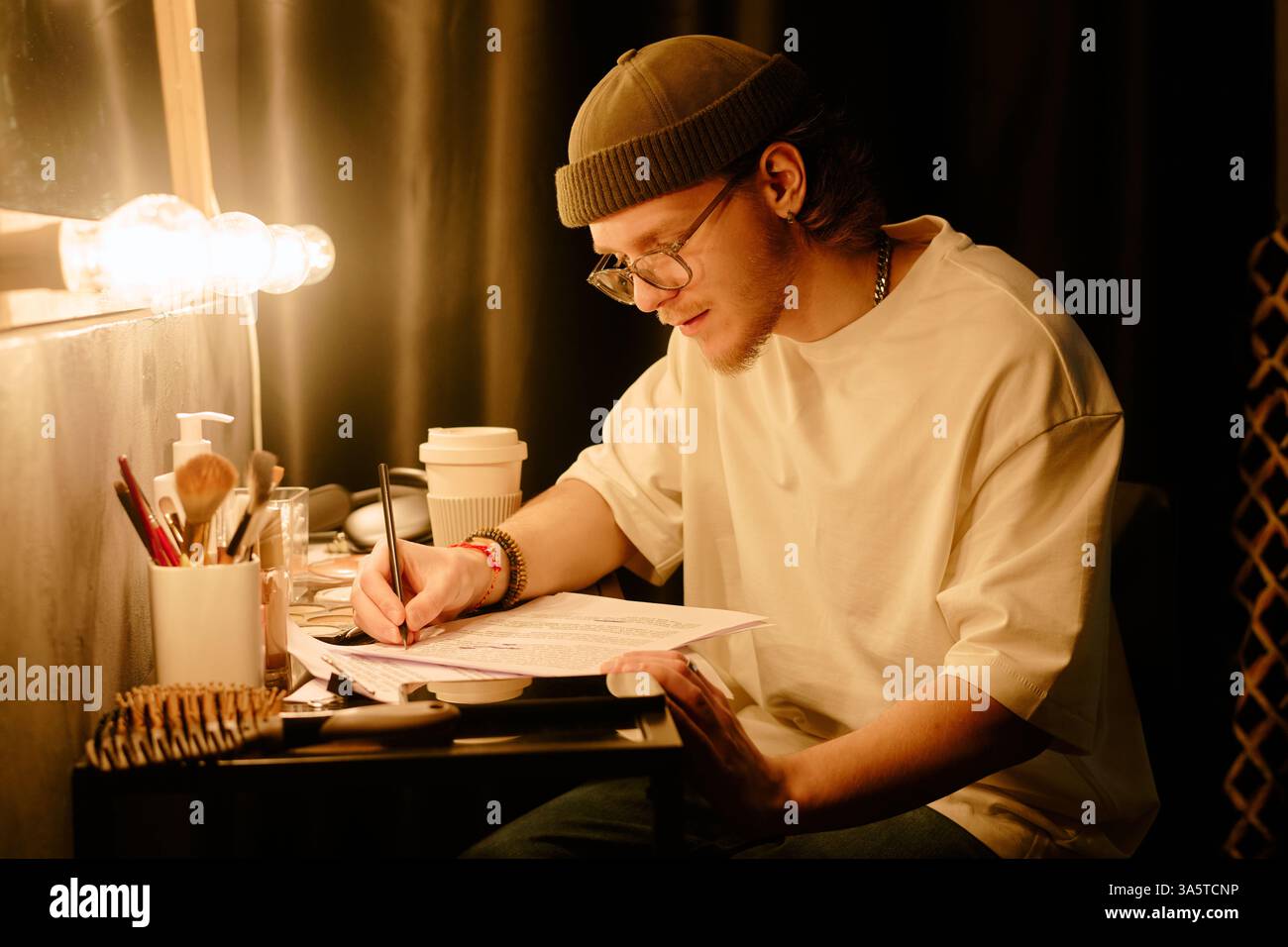 Young man focused on studying and writing notes at desk under warm lamp light, wearing beanie and glasses. Surrounded by study materials and personal items Stock Photo
