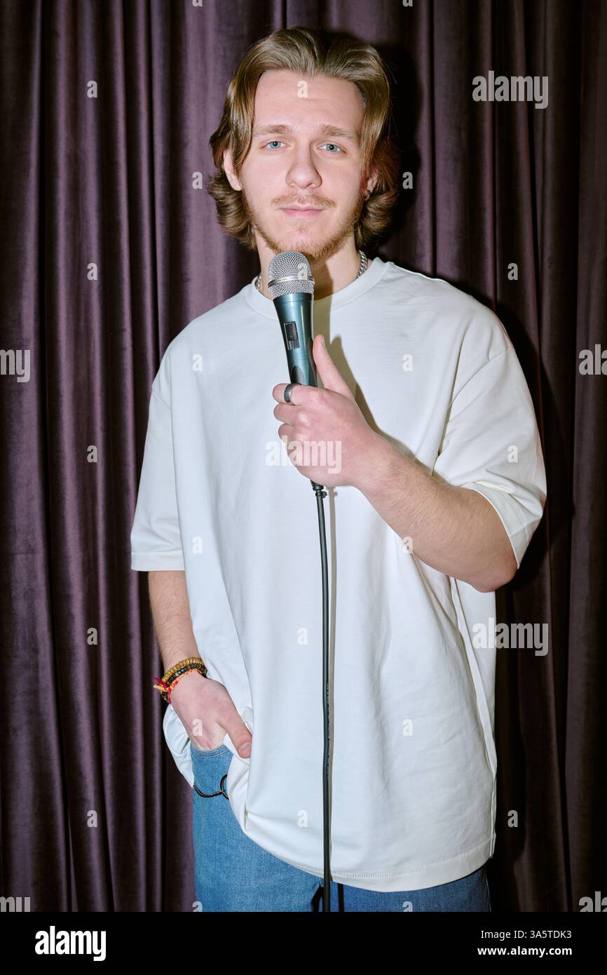 Portrait of young man with long hair standing in front of dark curtain, holding microphone, wearing casual clothes, smiling directly at camera, appearing confident Stock Photo