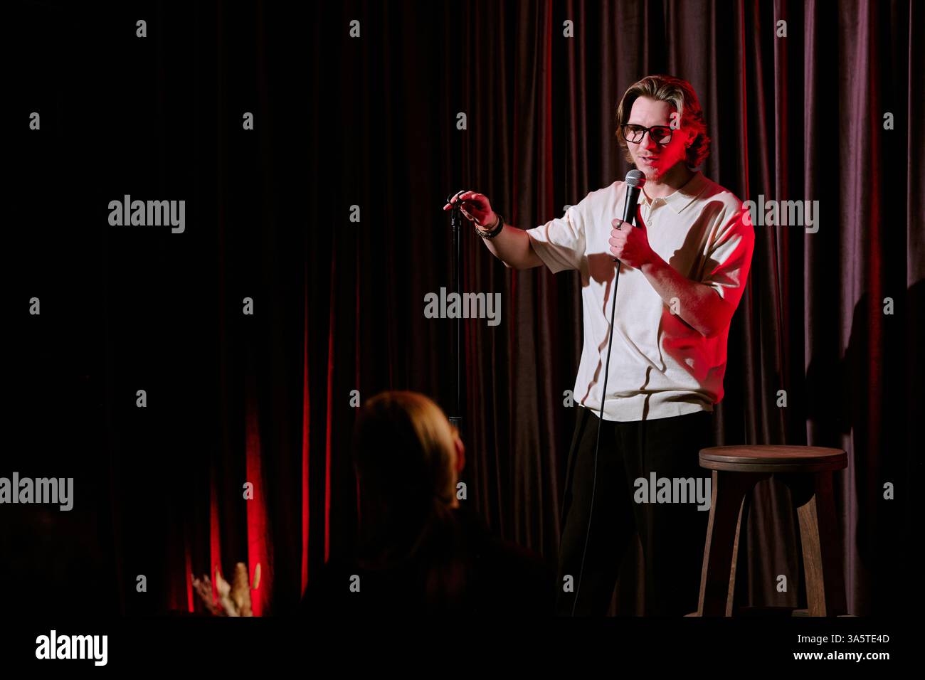 Comedian performing stand-up routine on stage, holding microphone and engaging with audience under dramatic lighting conditions Stock Photo
