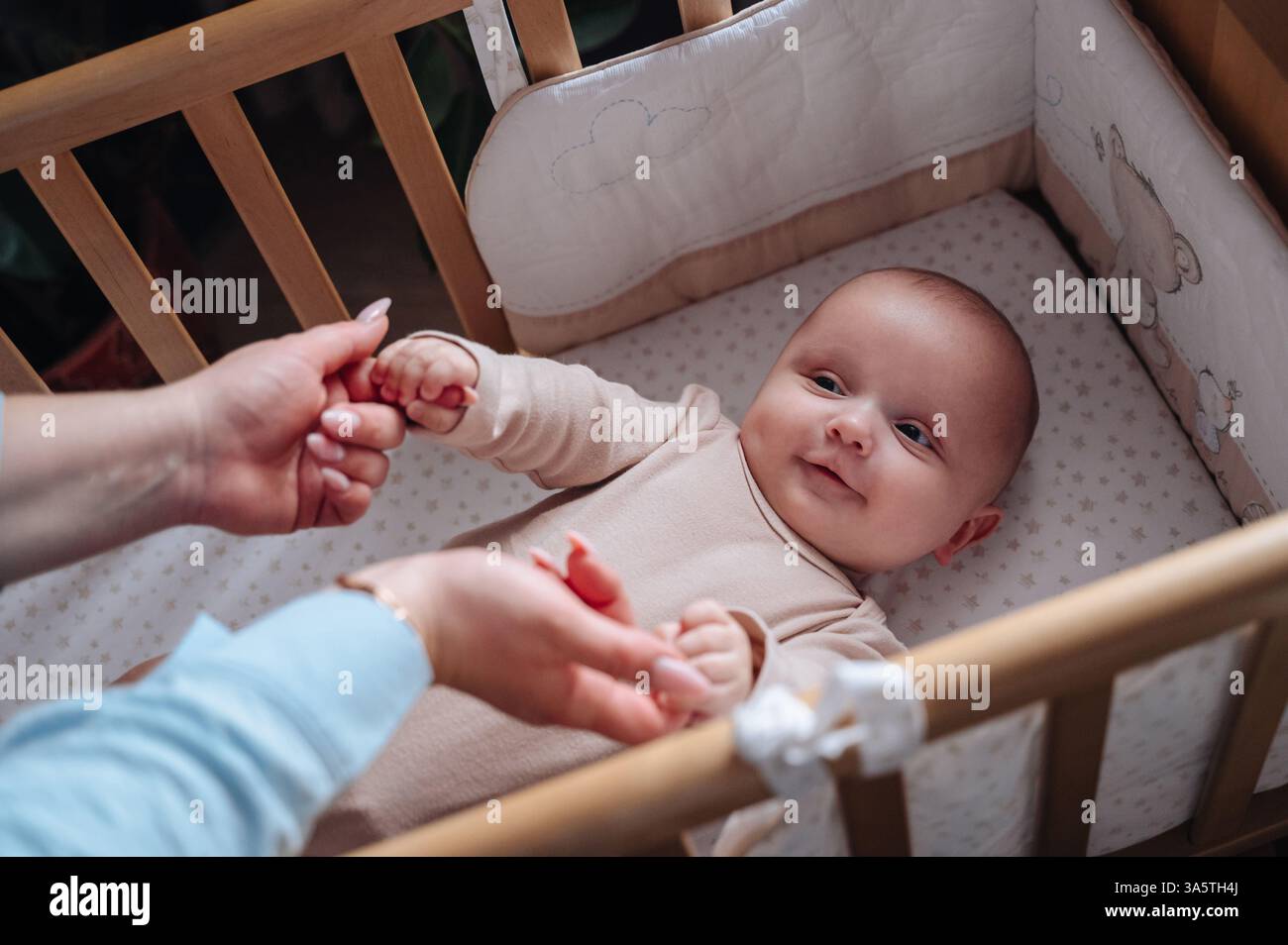 A loving mother gently holds the hands of her baby lying in a crib, creating a tender  Stock Photo