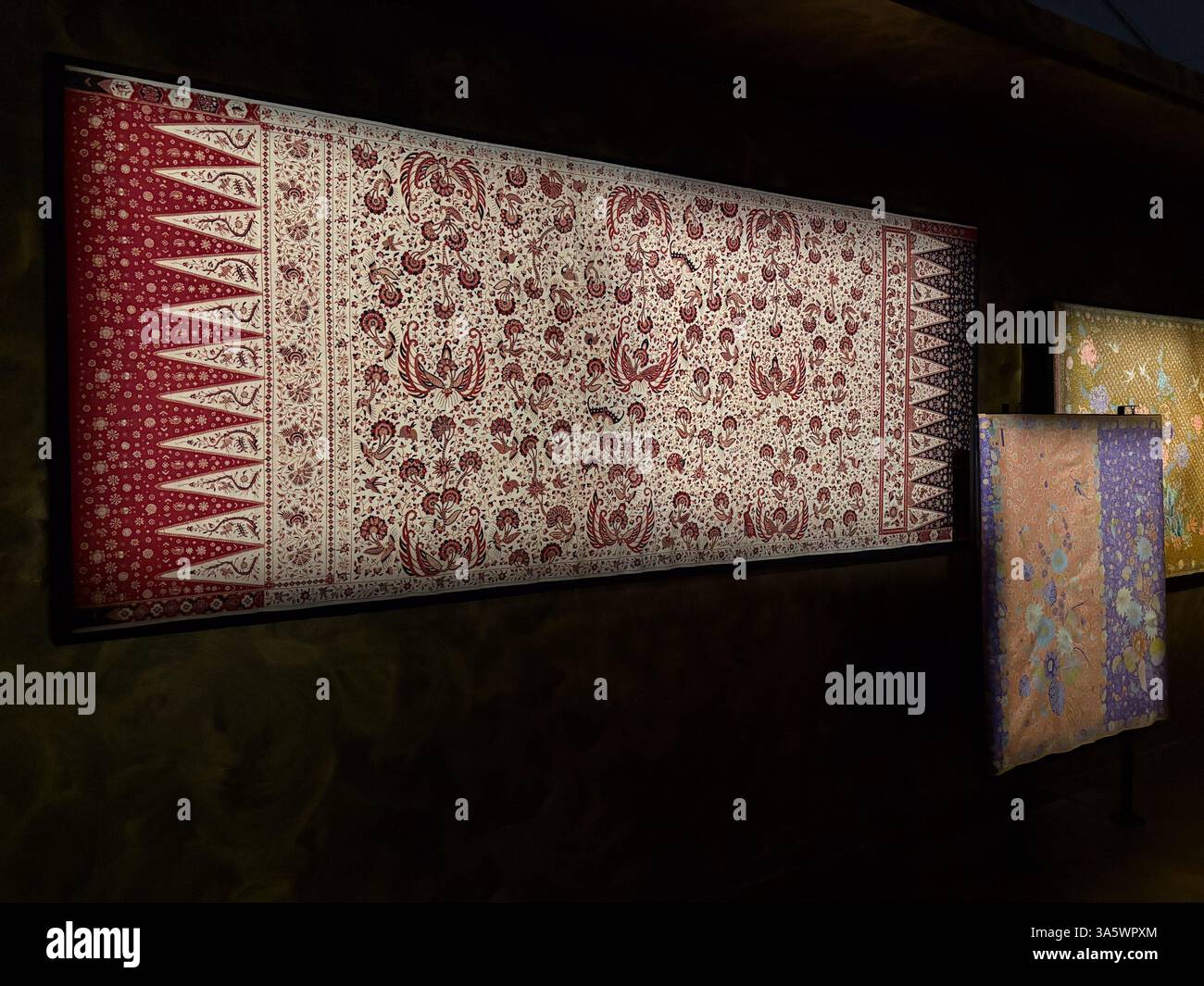 Display of a batik textile design with red and white patterns. Traditional Indonesian wax-resist dyeing technique on display at a museum. Stock Photo