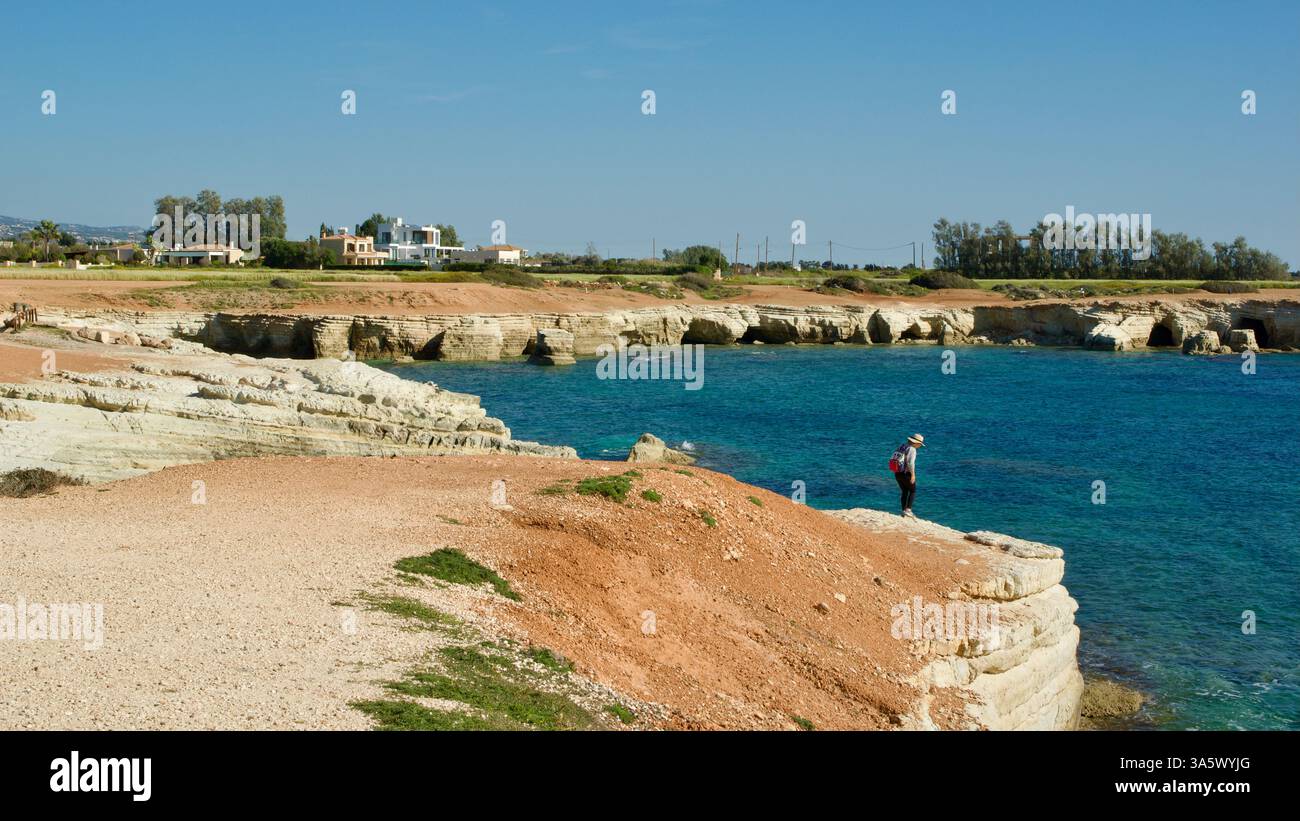 A scenic coastline featuring vivid blue waters, rocky shoreline, and a person gazing at the sea. Modern houses and green foliage decorate the distance Stock Photo