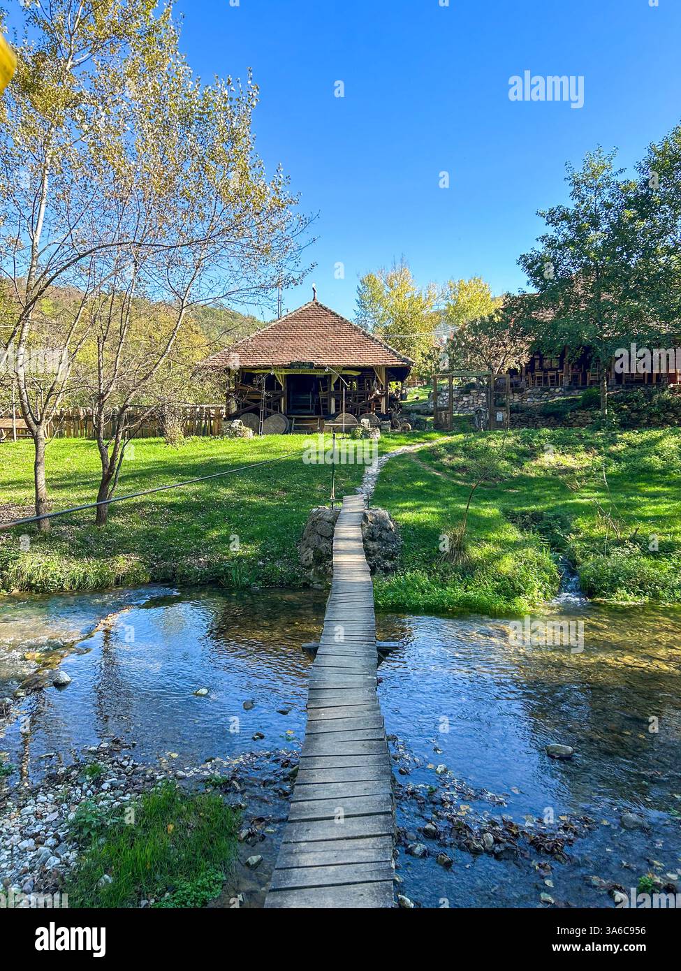 A wooden bridge stretches over the Gradac River, leading to a charming house surrounded by nature, creating a peaceful and picturesque scene. - Smartphone Captured Stock Image