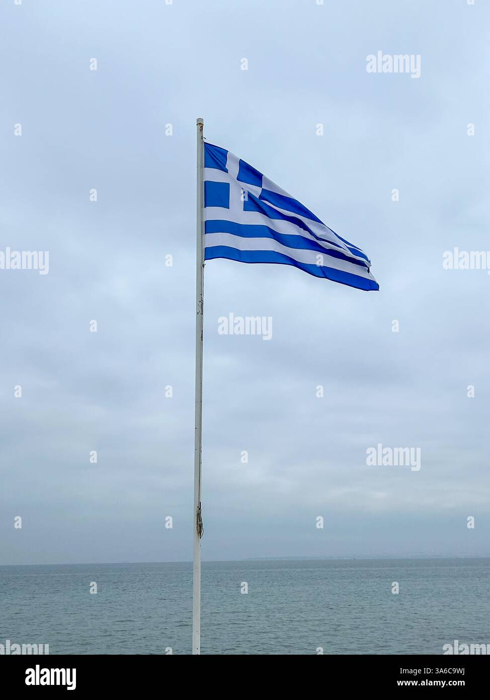 The Greek flag waves proudly by the sea under a cloudy sky, adding a touch of national spirit to the moody coastal atmosphere. - Smartphone Captured Stock Image