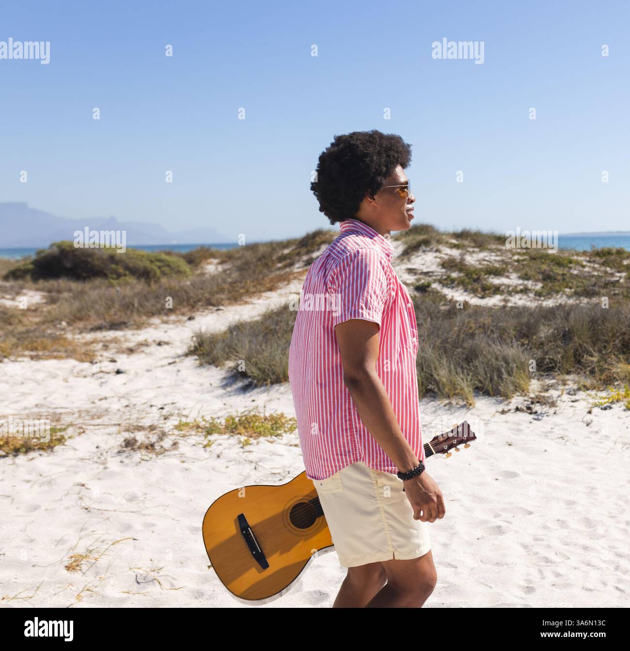 Young man walking on sandy beach carrying guitar, enjoying sunny day outdoors Stock Photo