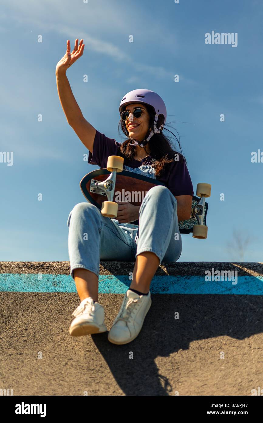 Young latin woman wearing helmet and sunglasses waving while sitting on a pump track ramp holding a surfskate, enjoying a sunny day practicing her fav Stock Photo