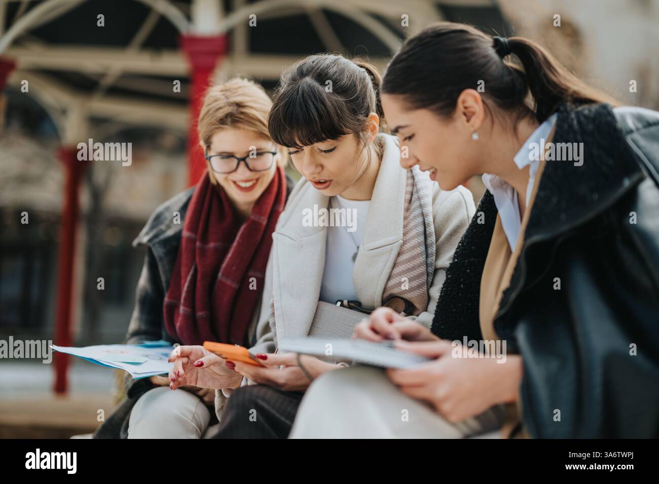 Three businesswomen collaborating outdoors on work materials and a mobile device Stock Photo