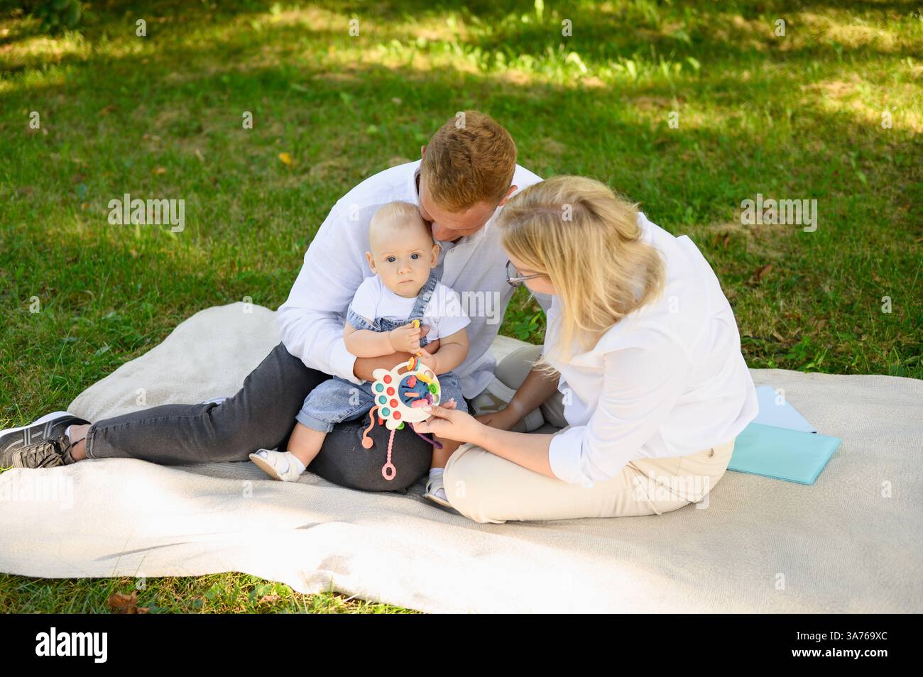 Man and woman sit comfortably on blanket spread on grass, enjoying picnic on sunny day with child on lap. Child plays with educational toy during walk Stock Photo