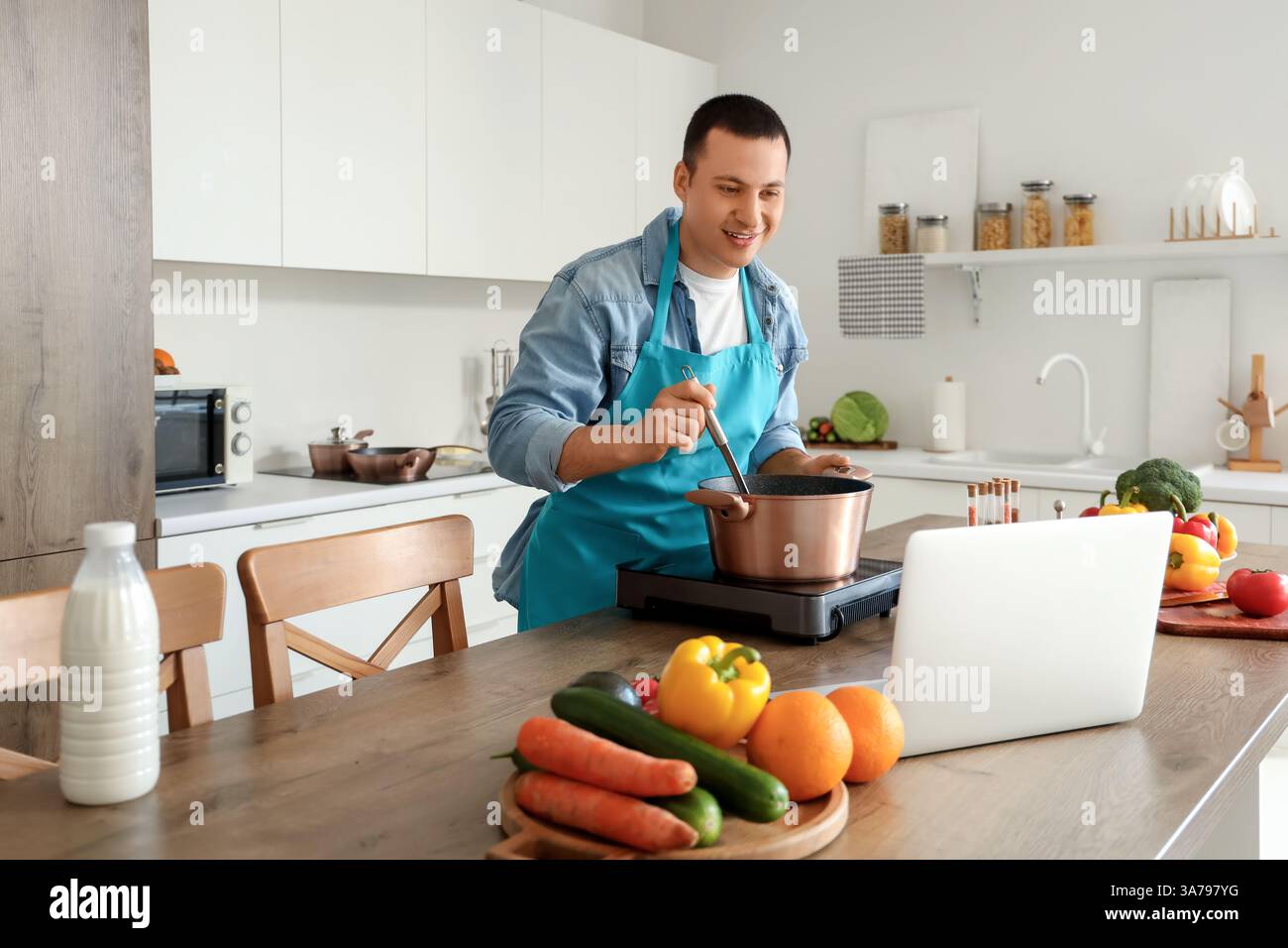 Young man with laptop cooking soup in kitchen Stock Photo