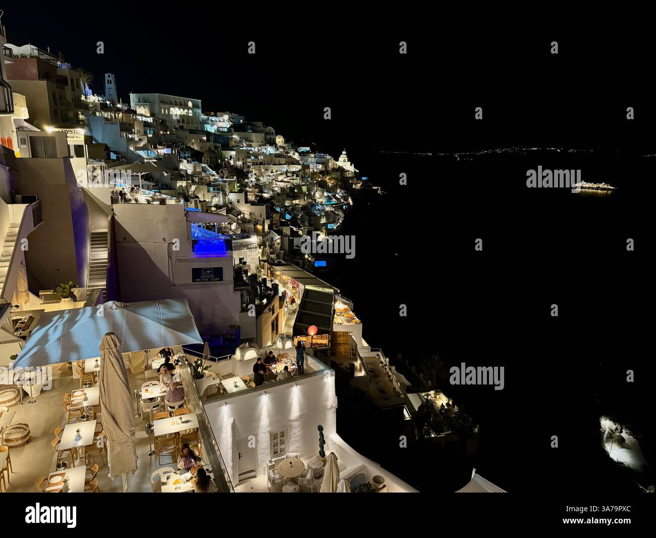 A night-time view of the steep slopes of Santorini, with tourists enjoying dinner on the illuminated terraces of multiple restaurants and hotels. - Smartphone Captured Stock Image