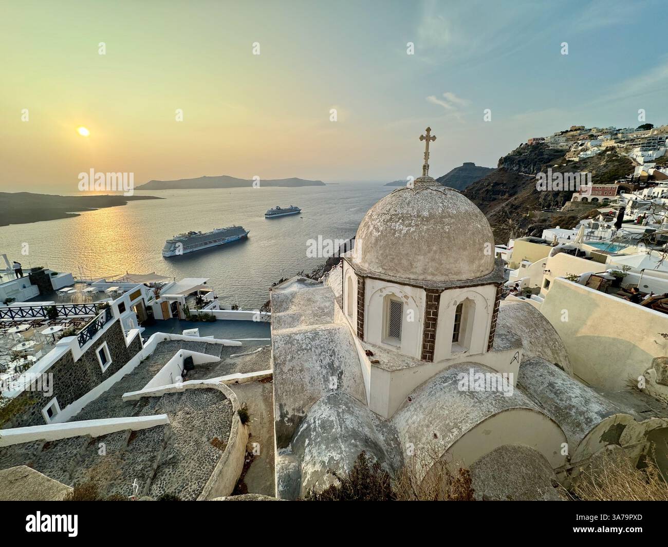 A view overlooking the dome of The Church of Saint John the Theologian in Santorini at sunset, with two large cruise ships at anchor in the caldera. - Smartphone Captured Stock Image