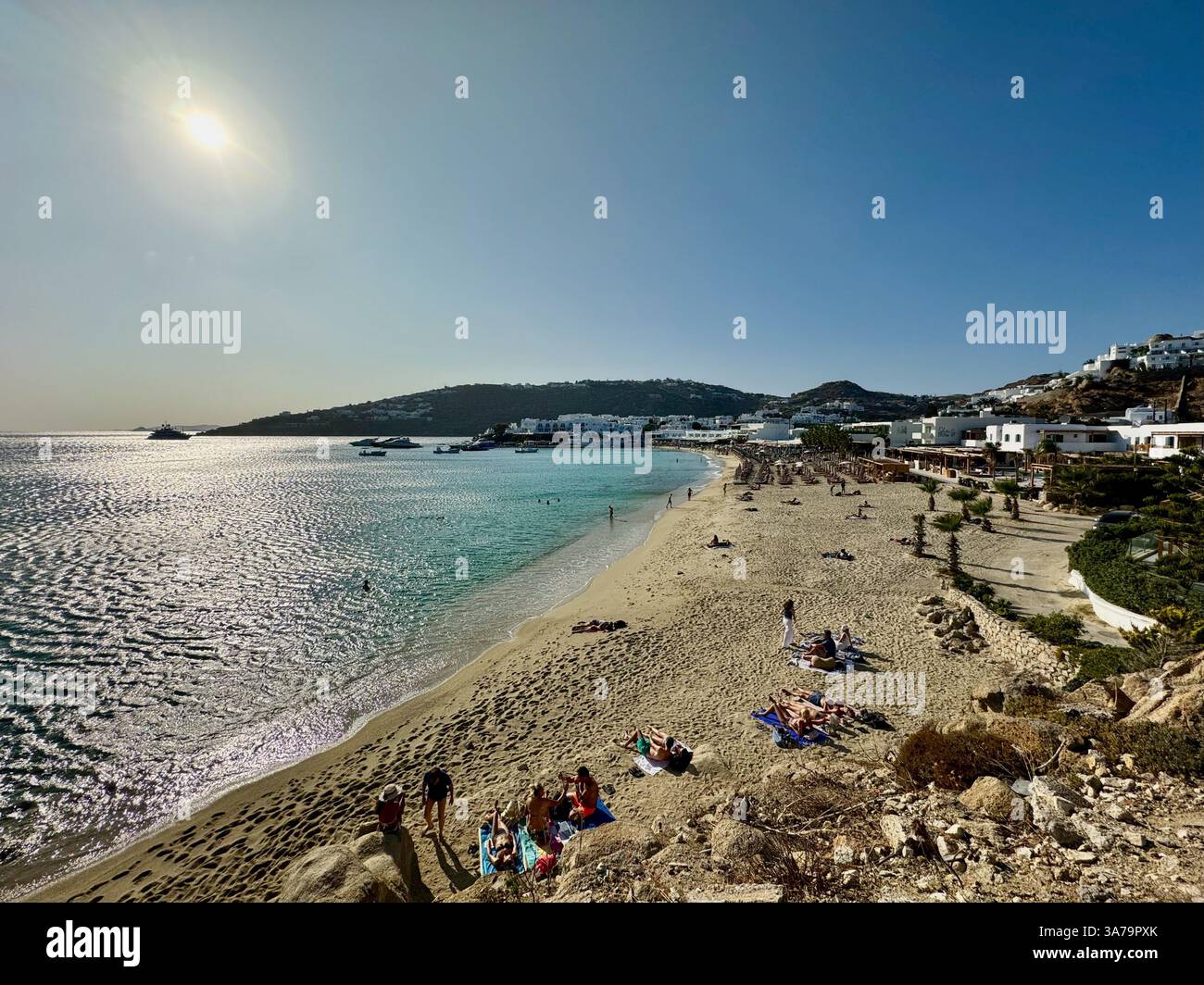 Sunbathers enjoy the late afternoon sunshine at Platis Gialos beach on Mykonos Island, Greece, with Branco Beach Club visible in the distance. - Smartphone Captured Stock Image
