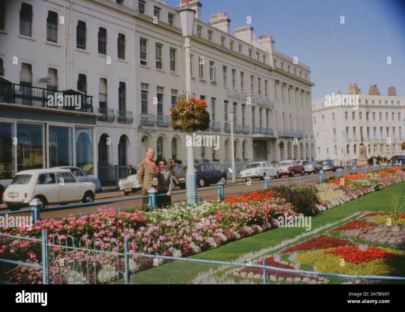 A couple pose by the colourful Carpet Gardens in Eastbourne, UK, on a sunny day in the 1960s. Captured on 35mm slide film, the photo shows the vibrant flower beds, classic cars, and the iconic Claremont Hotel along the seafront. Stock Photo