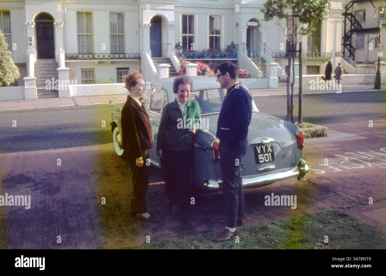 Three people gather by a Riley 1.5 saloon car parked on a residential street in Eastbourne, England during the 1960s. This classic British vehicle, sharing its design roots with the Wolseley 1500, reflects the postwar automotive style of the time. Stock Photo