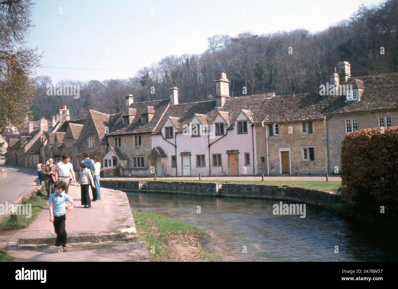 A picturesque view of Castle Combe in the 1960s, a quintessential English village nestled in the Cotswolds. The image captures rows of charming honey-coloured stone cottages lining the stream, with families leisurely strolling by on a sunny day. Stock Photo