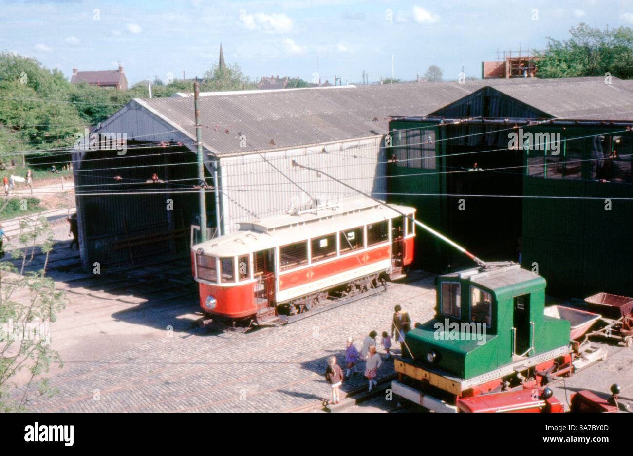 A 1960s scene at Crich Tramway Museum in Derbyshire, captured on 35mm slide film. A restored red and cream tram sits on cobbled tracks outside a corrugated metal depot, with families and children nearby enjoying a day at the museum. Overhead wires and vintage industrial details enhance the historic charm. Stock Photo