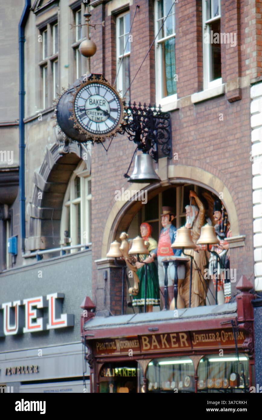 The Baker’s clock in Gloucester, England, photographed in 1974, features life-size automaton figures that strike the bells beneath a traditional Greenwich Time clock face. Located above the Baker jeweller and optician’s shop, this whimsical mechanical display is a well-known local landmark and a fine example of decorative street horology. Stock Photo