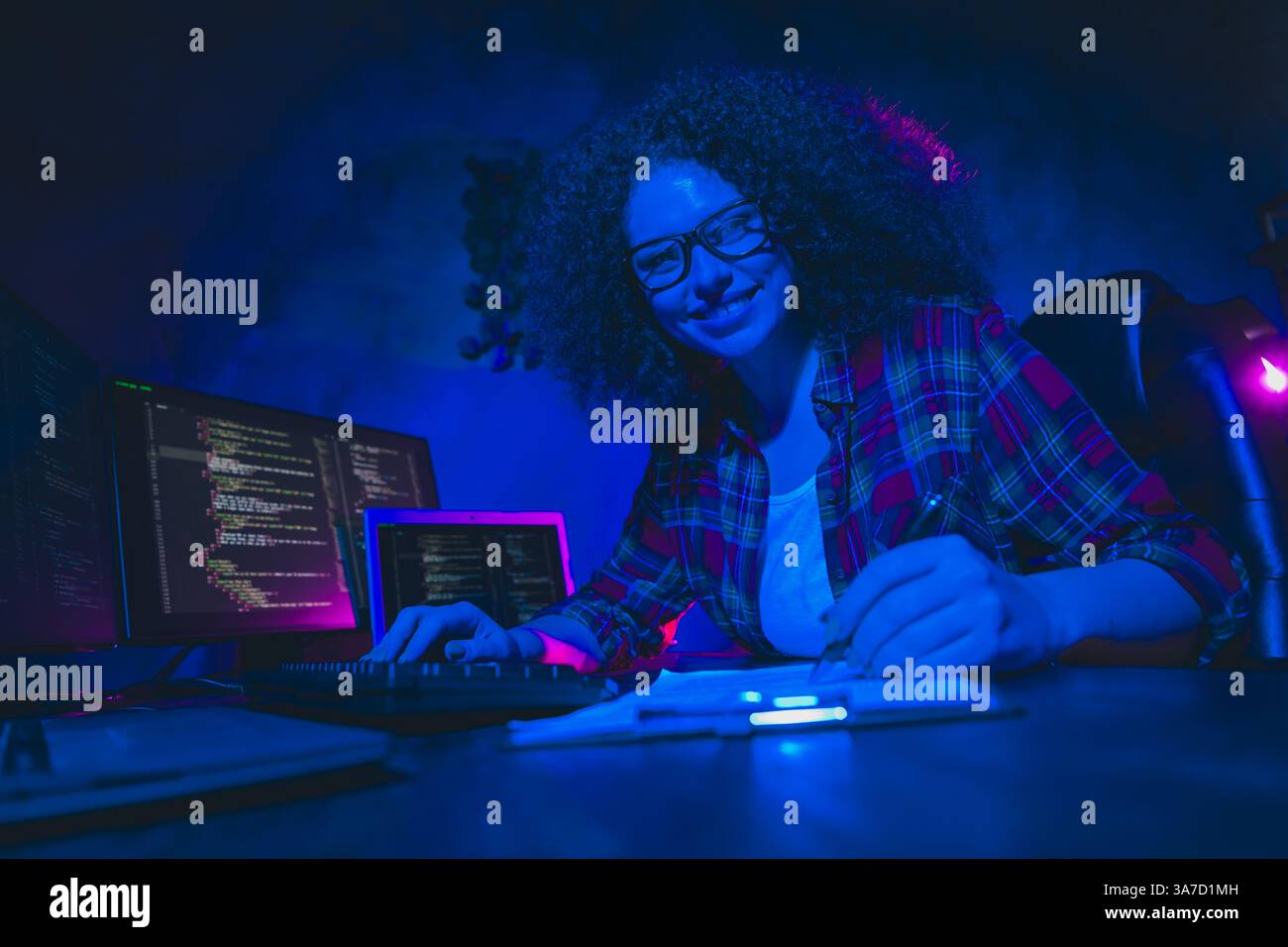 Young female programmer working on code in a dimly lit workspace, surrounded by multiple screens Stock Photo