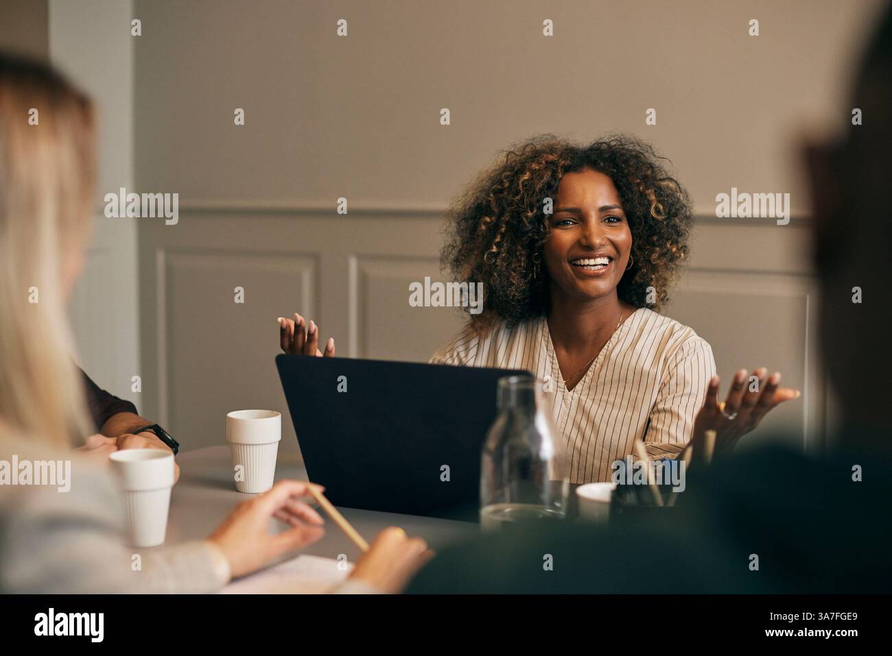Happy curly haired young businesswoman sitting with laptop while discussing during meeting in office Stock Photo