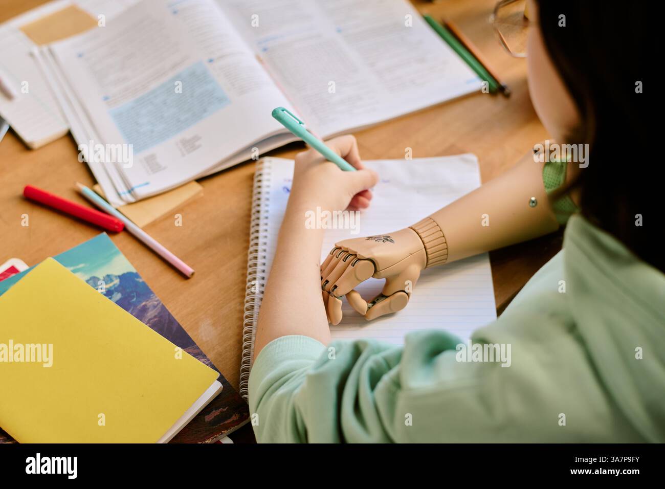 Person using prosthetic hand while writing at desk cluttered with books, notebooks, and stationery. Focused studying scene suggesting determination and perseverance Stock Photo