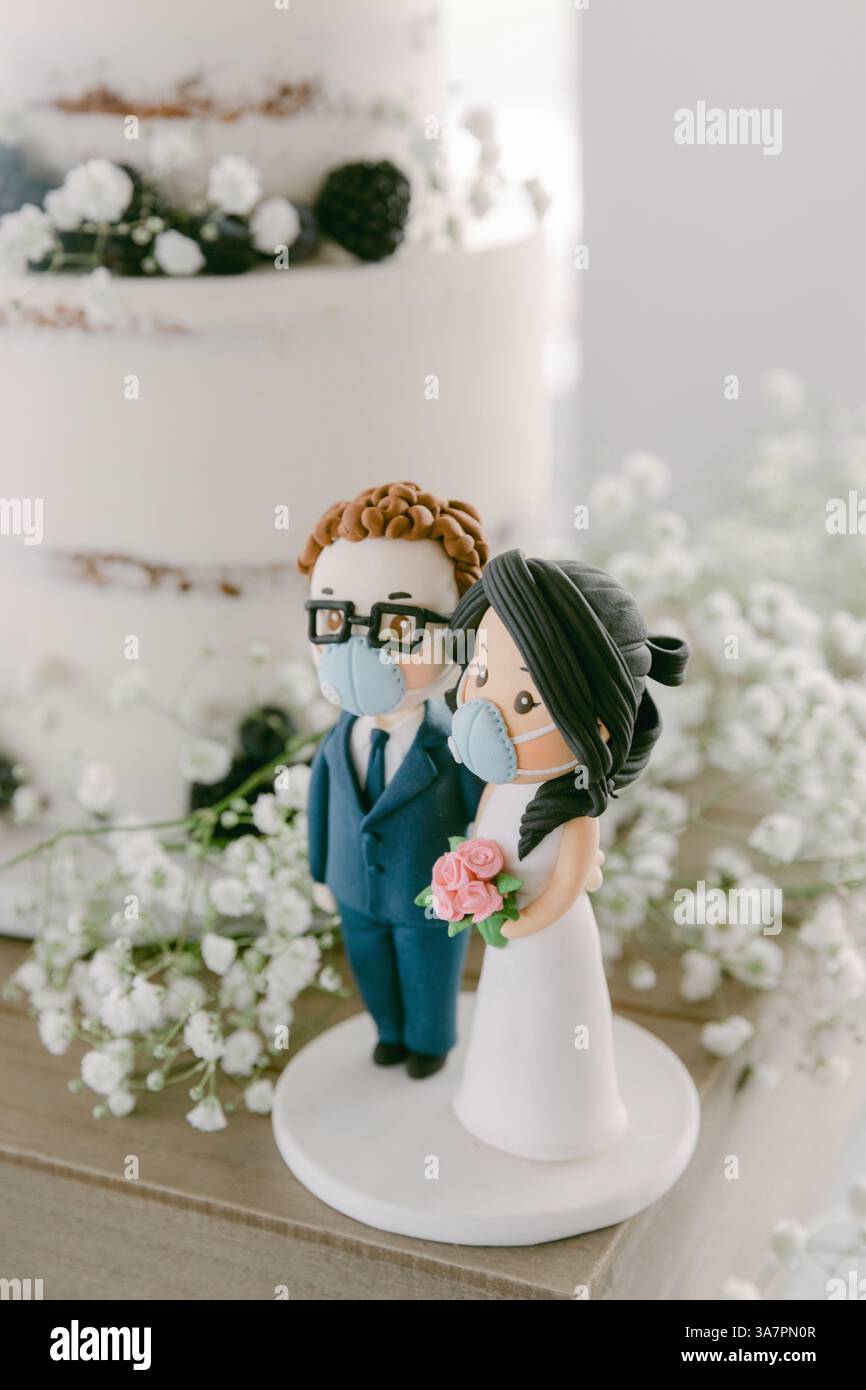 A couple stands side by side, wearing masks while holding flowers, close to their wedding cake decorated with flowers. Stock Photo