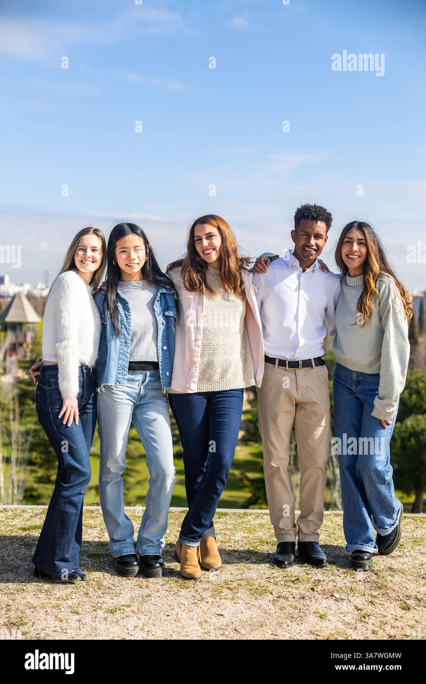 Five multi ethnic students standing together in a park with arms around each other, smiling Stock Photo