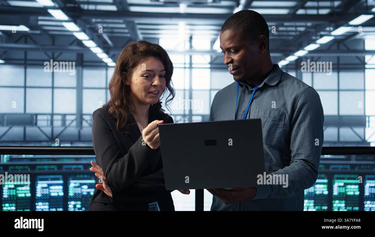 Engineers using laptop in data center, managing server virtualization, running scripts. IT specialists using programming language on notebook in server hub, finetuning network protocols, camera B Stock Photo