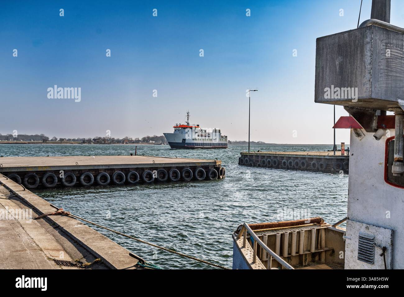 A scenic view of a ferry arriving at a harbor under a bright blue sky, surrounded by calm waters. The environment includes dock structures and portray Stock Photo