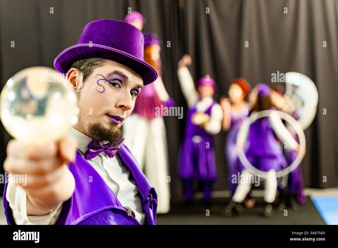 Magician wearing purple top hat and suit showing crystal ball with circus performers juggling and dancing in background Stock Photo