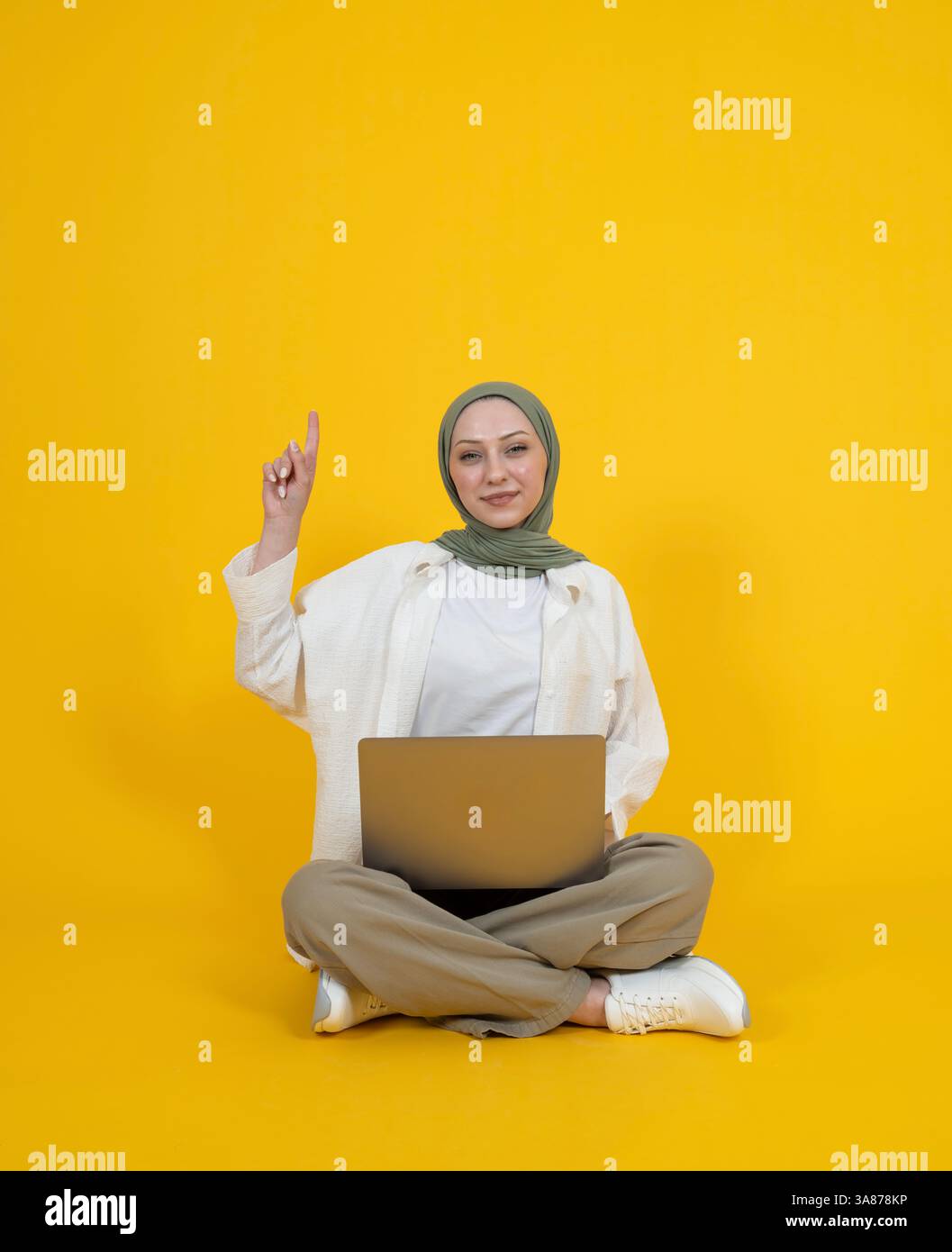Caucasian muslim girl points above. Girl wear abaya hijab sit floor laptop pc computer on her lap. I have a great idea concept idea image. Yellow. Stock Photo