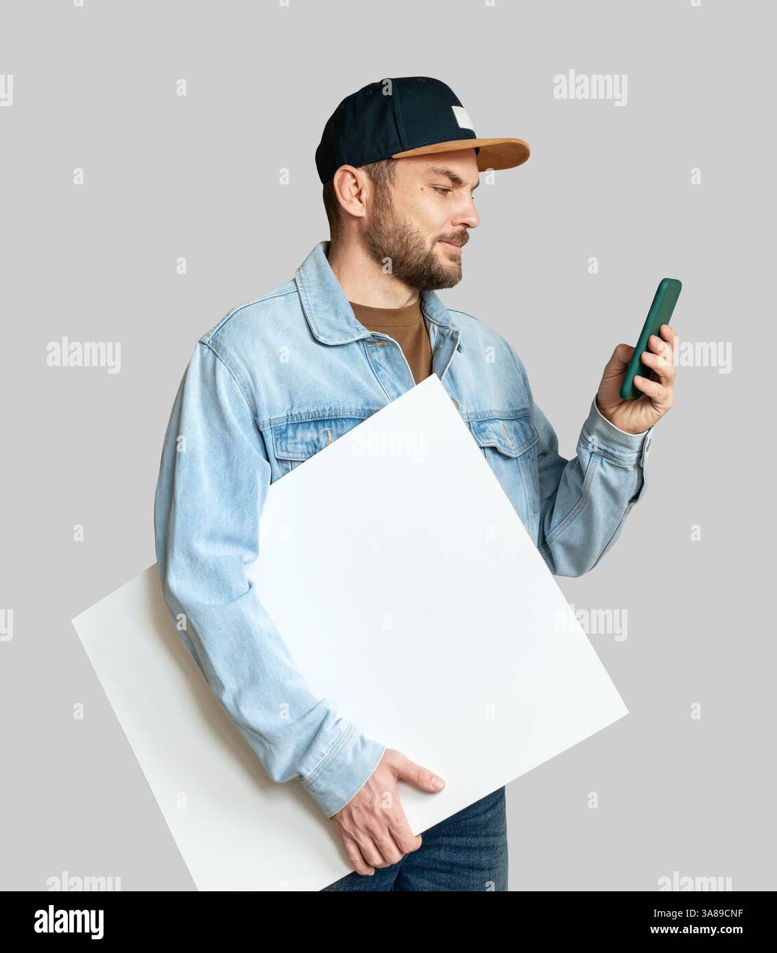 Man in denim jacket and snapback holding blank white board while using smartphone Stock Photo