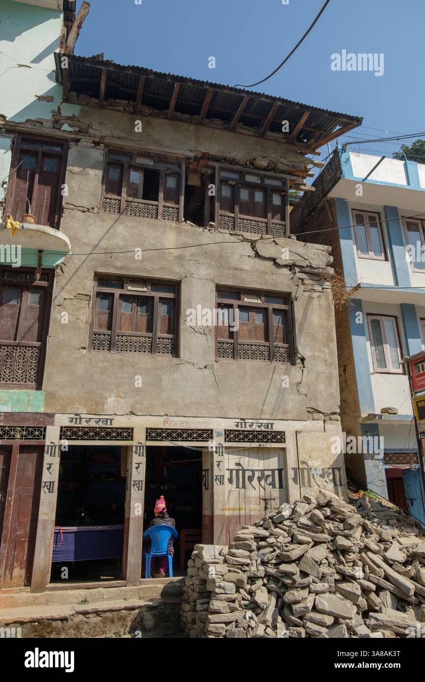 A person sits in the doorway of a building severely damaged by an earthquake in Nepal, with debris piled outside. Stock Photo