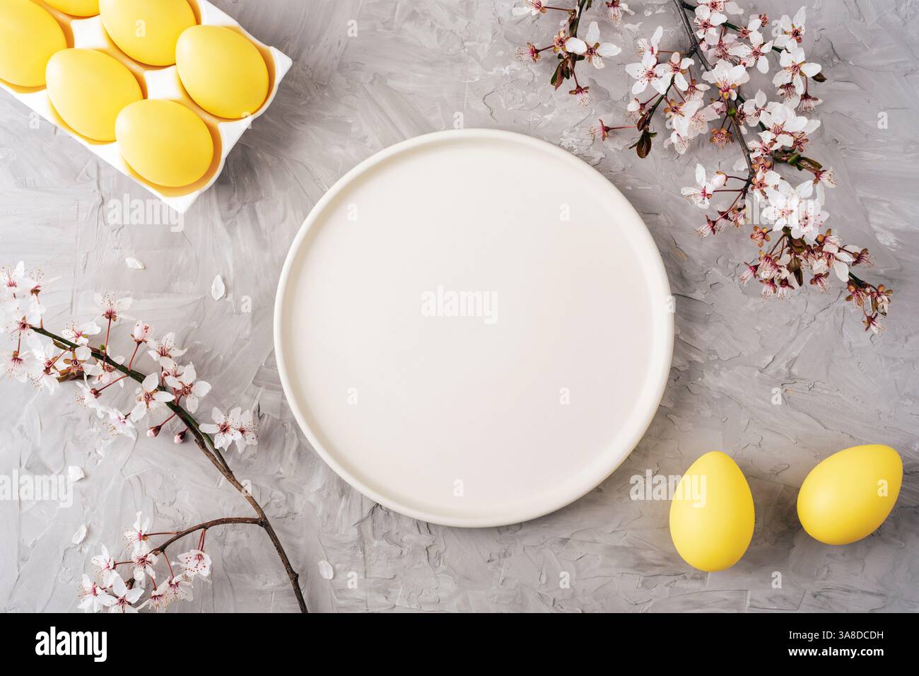 Yellow Easter eggs on gray background with empty plate and cherry blossom branches. Top view, flat lay, mockup. Stock Photo