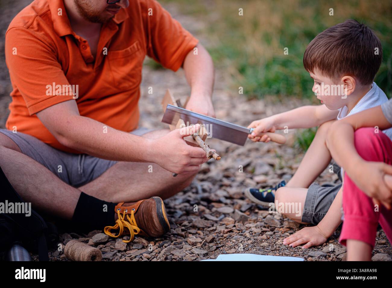 A father guides his young son in using a saw to cut a small branch. The outdoor setting suggests a hands-on learning experience, camping, or bushcraft Stock Photo