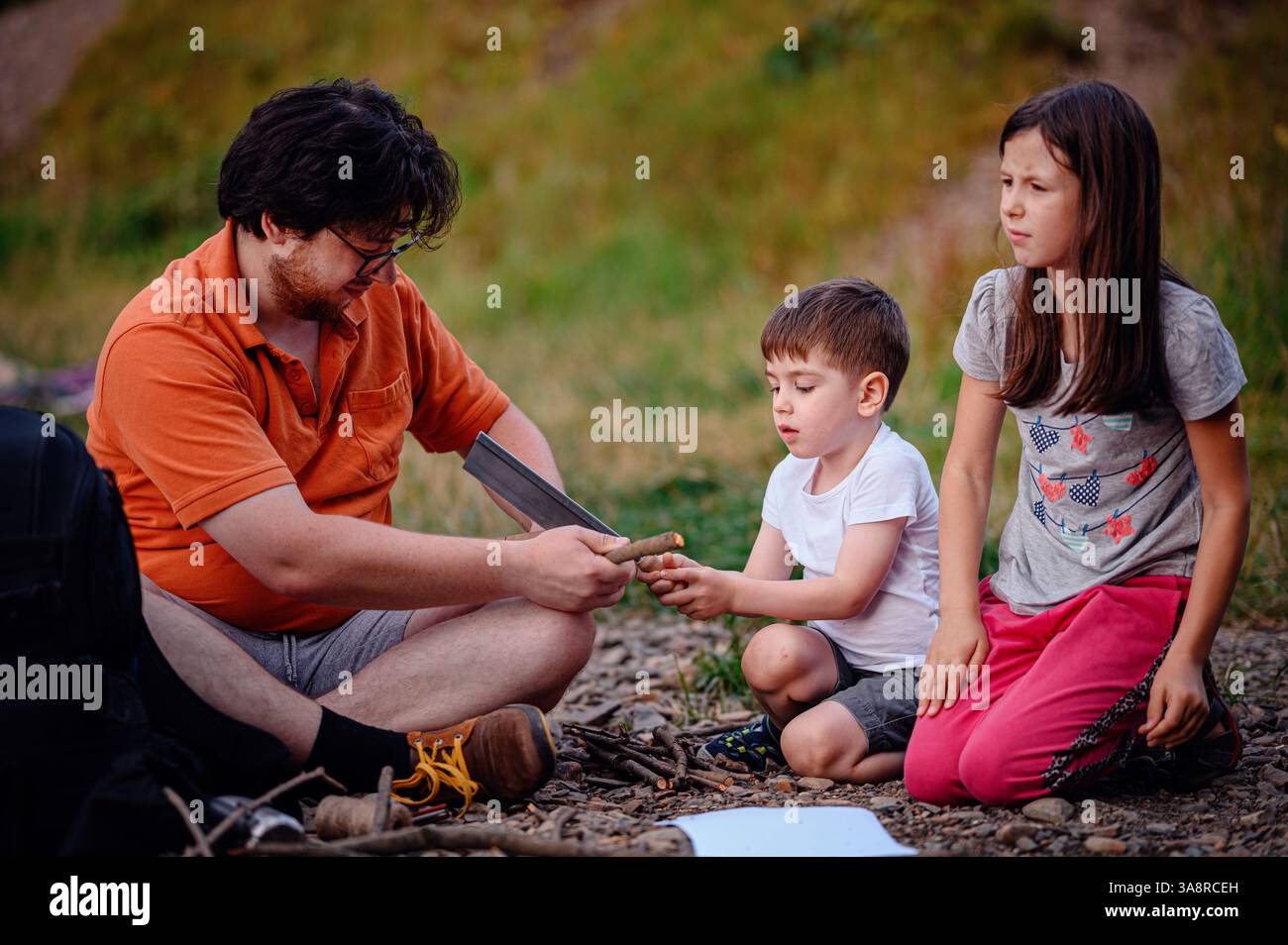 A father teaches his children how to use a saw and gather sticks during an outdoor activity. A hands-on learning experience promoting teamwork, skill- Stock Photo
