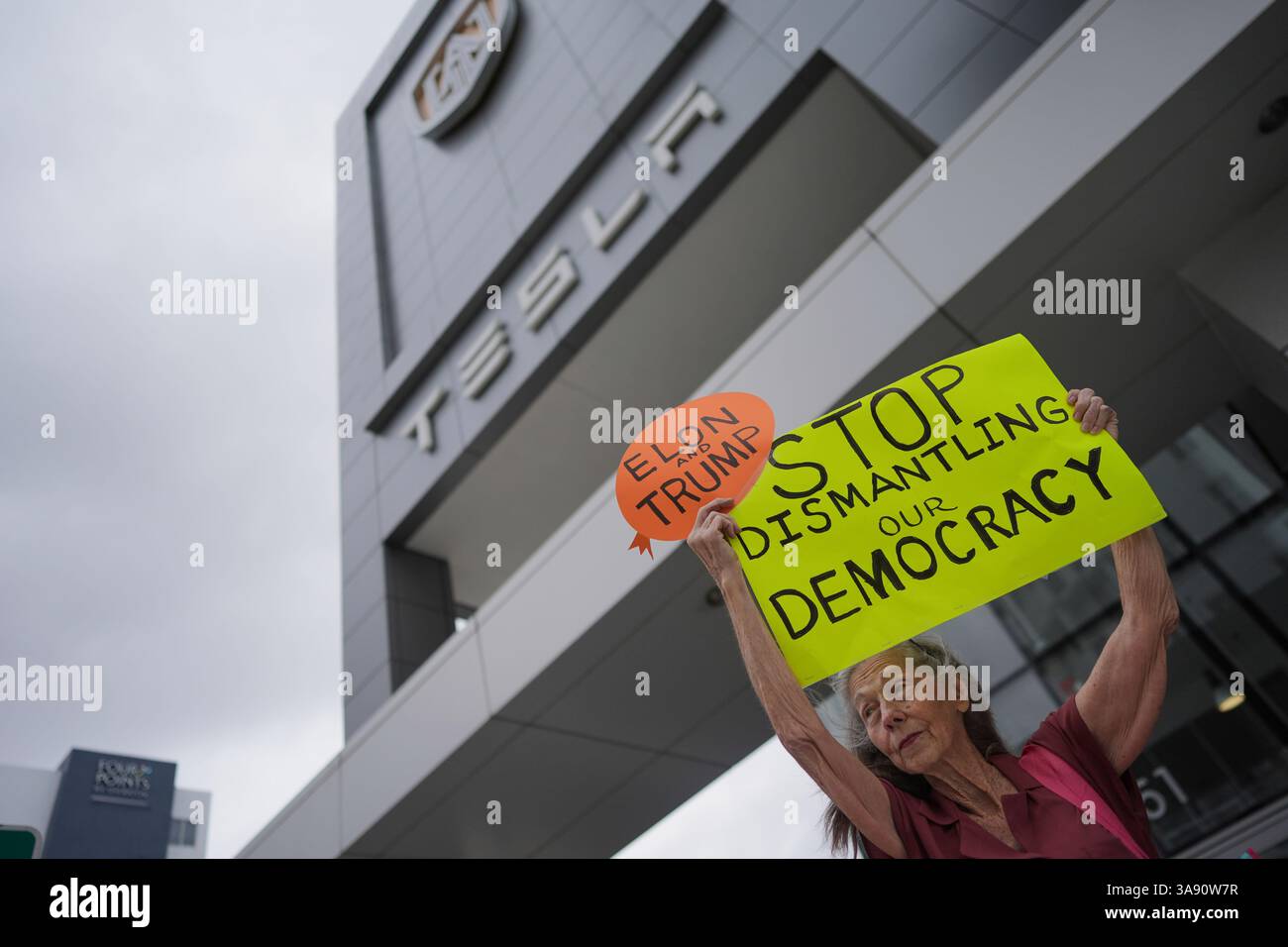 A woman holds up a sign as people protest in front of a Tesla ...