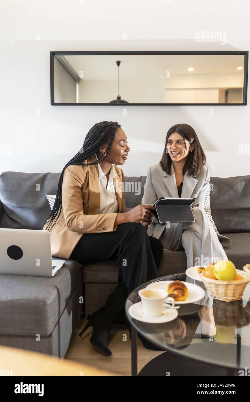 Two businesswomen are sitting on a sofa in a hotel room, using a tablet and laptop during a productive meeting, enjoying coffee and croissants Stock Photo