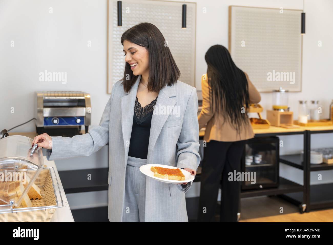 Businesswoman smiling while choosing food from breakfast buffet in hotel during business trip, enjoying a slice of toasted bread with jam Stock Photo