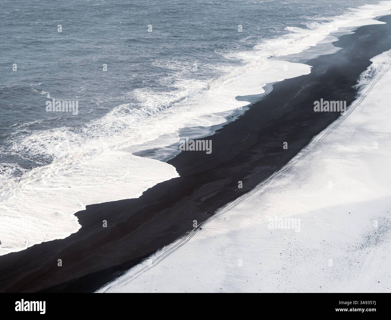Aerial view of Dyrhólaey black beach in Iceland, where waves meet volcanic sand and fresh snow. Abstract winter coastal landscape with bold contrast a Stock Photo
