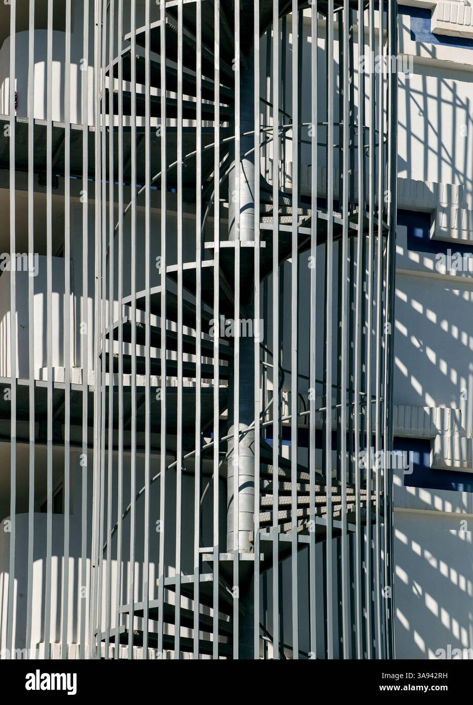 An emergency spiral staircase enclosed by vertical metal bars provides a secure fire escape, with striking architectural patterns of shadows and lines. Stock Photo